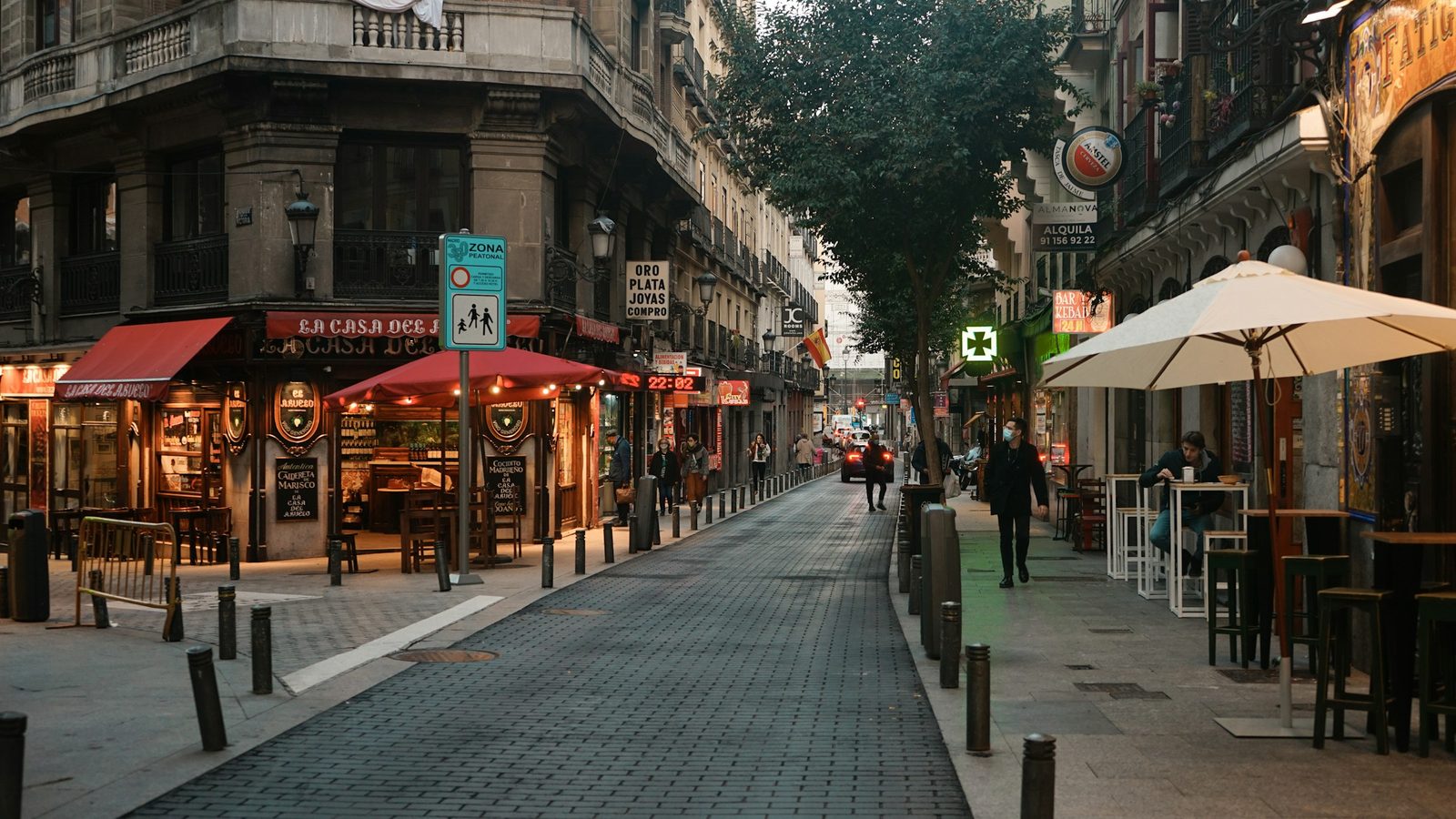 A pedestrian street in Madrid's Barrio de las Letras with small family restaurants and shops, red awnings, a café terrace with a white parasol and a few people walking at dusk