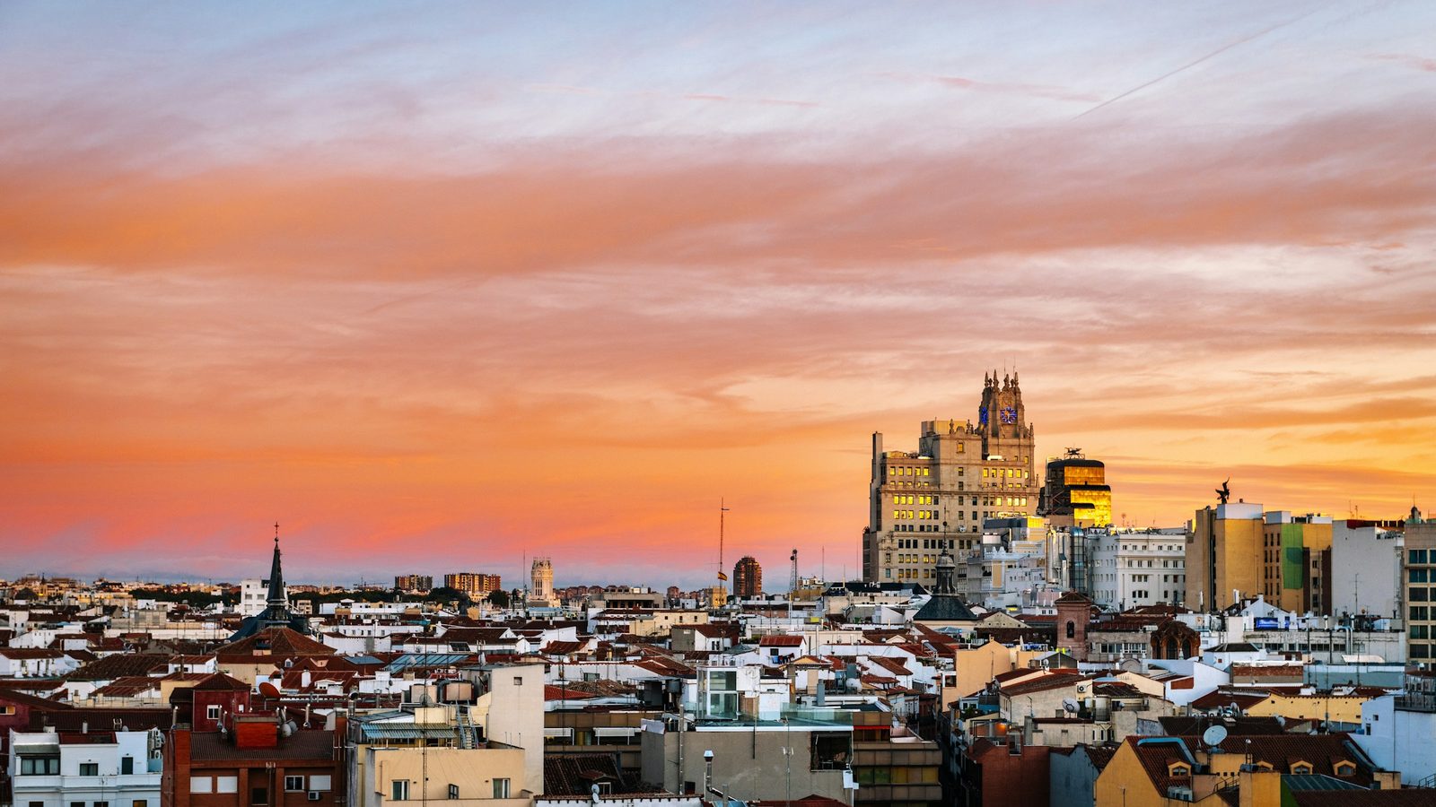 Madrid rooftops at sunset under an orange and pink sky, with the Telefónica Building's clock tower on Gran Vía rising above the city centre