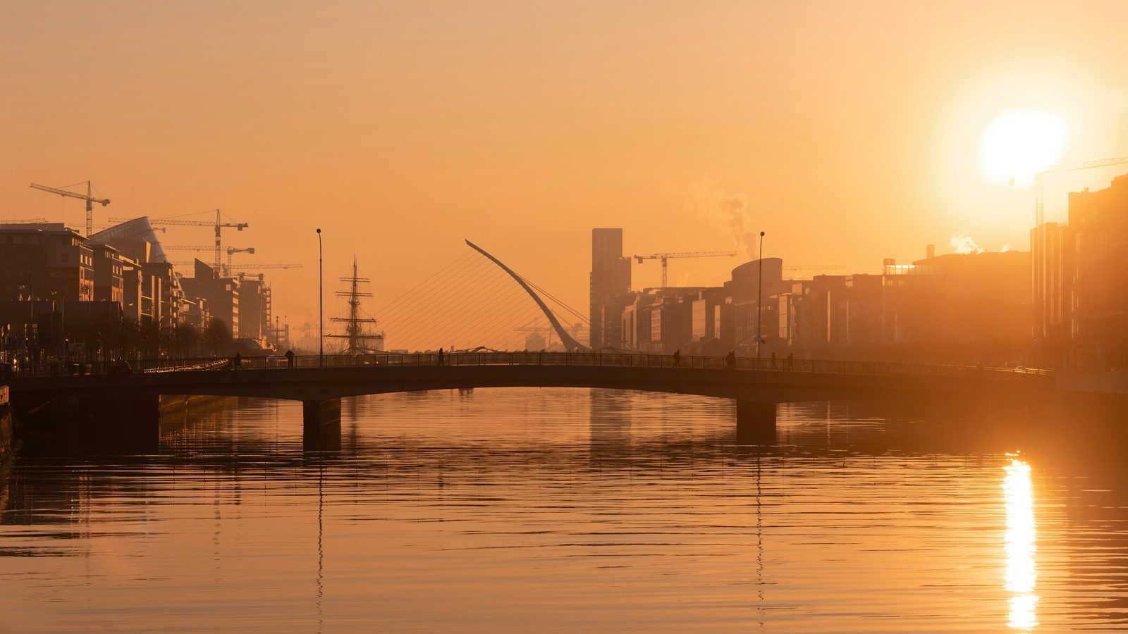 The River Liffey in Dublin at sunrise, with a footbridge, the harp-shaped Samuel Beckett Bridge and the Docklands skyline in orange light