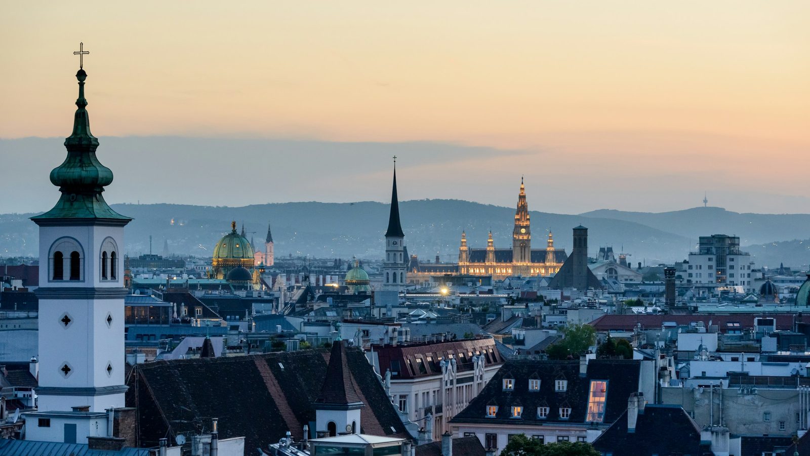 Vienna rooftops at dusk, with church towers, the lit-up City Hall and the hills of the Vienna Woods behind