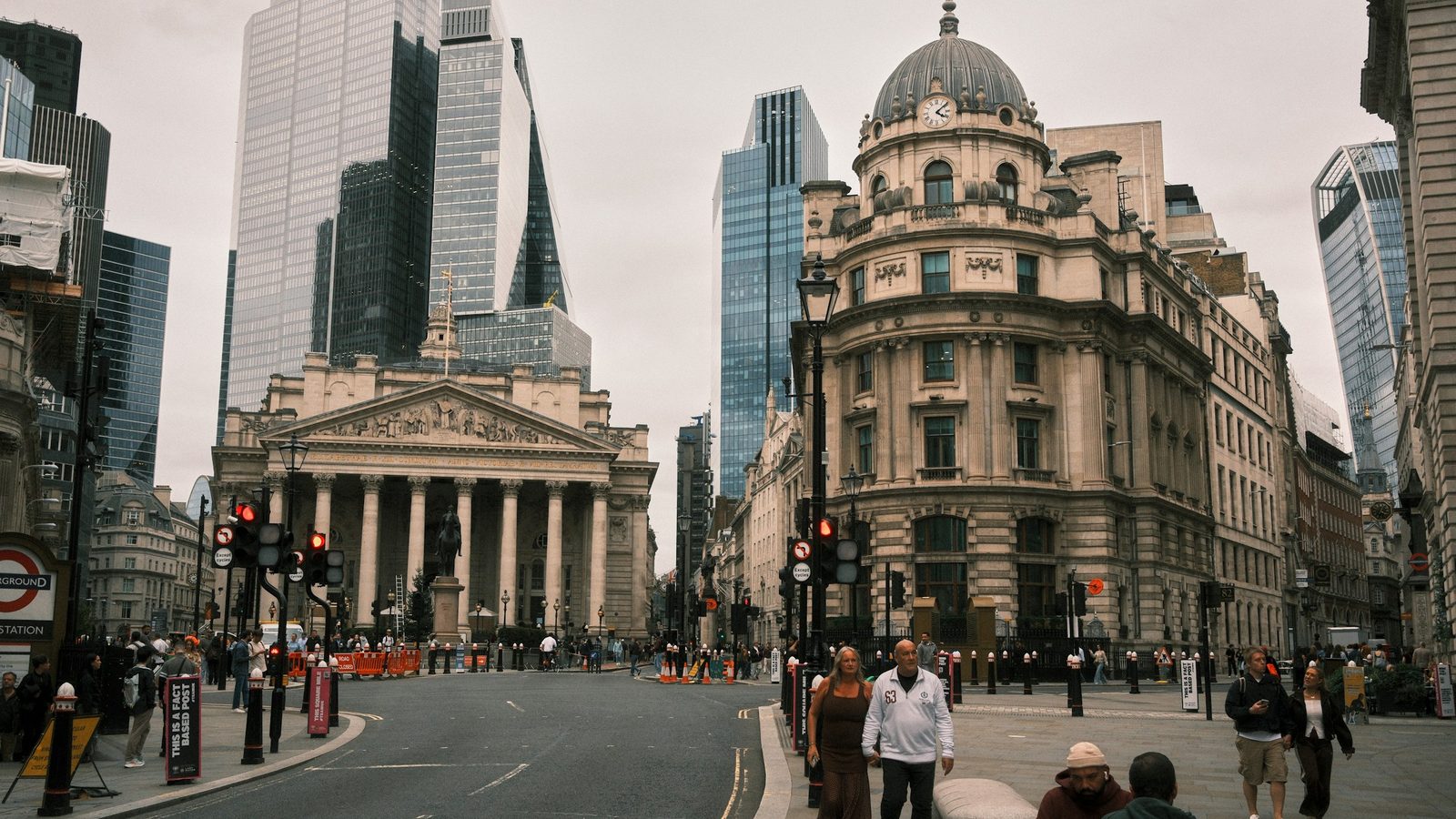 Pedestrians on the street at Bank junction in the City of London, with the columns of the Royal Exchange, a domed stone office building and glass skyscrapers behind them under a grey sky