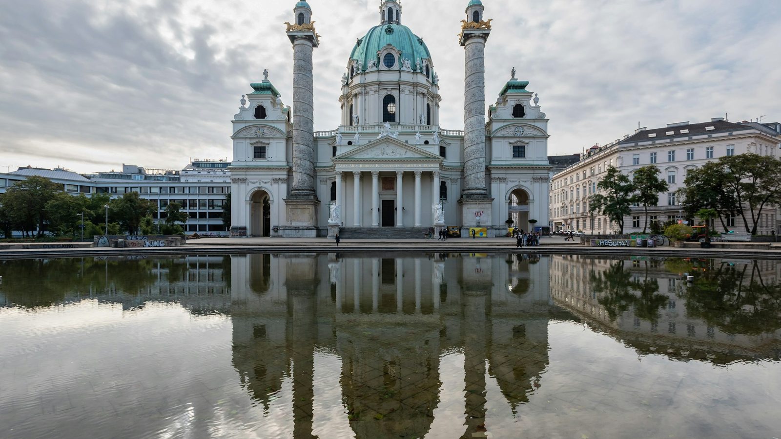 Karlskirche in Vienna, a white baroque church with a green dome and two carved columns, reflected in the pond on Karlsplatz under a cloudy sky