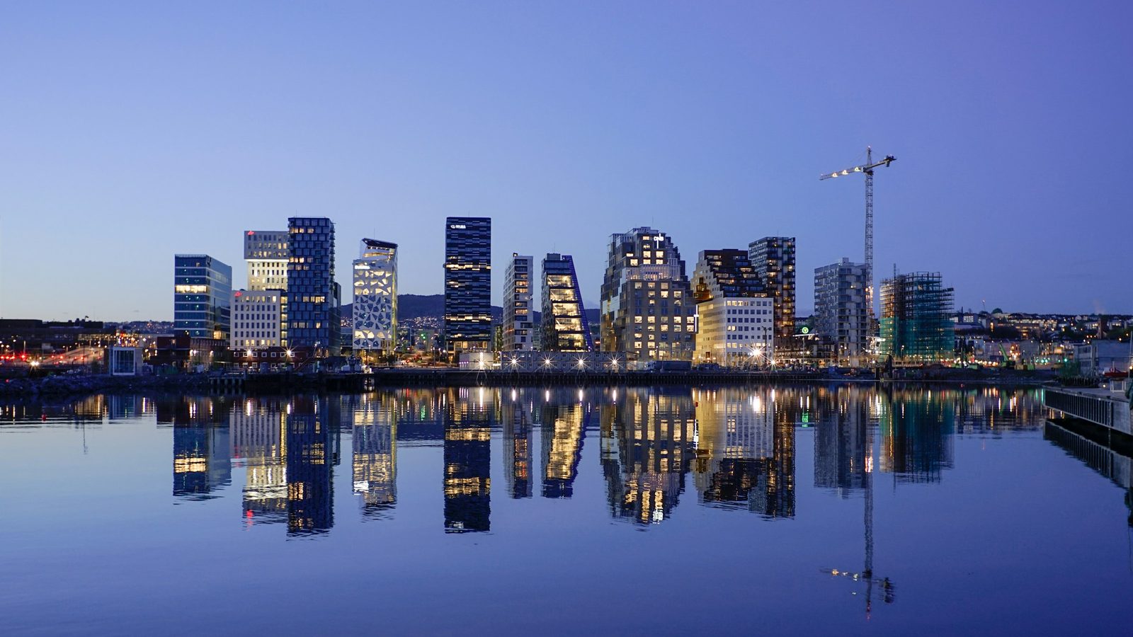The lit-up Barcode office towers in Bjørvika, Oslo, reflected in the still water of the fjord at dusk, with a construction crane
