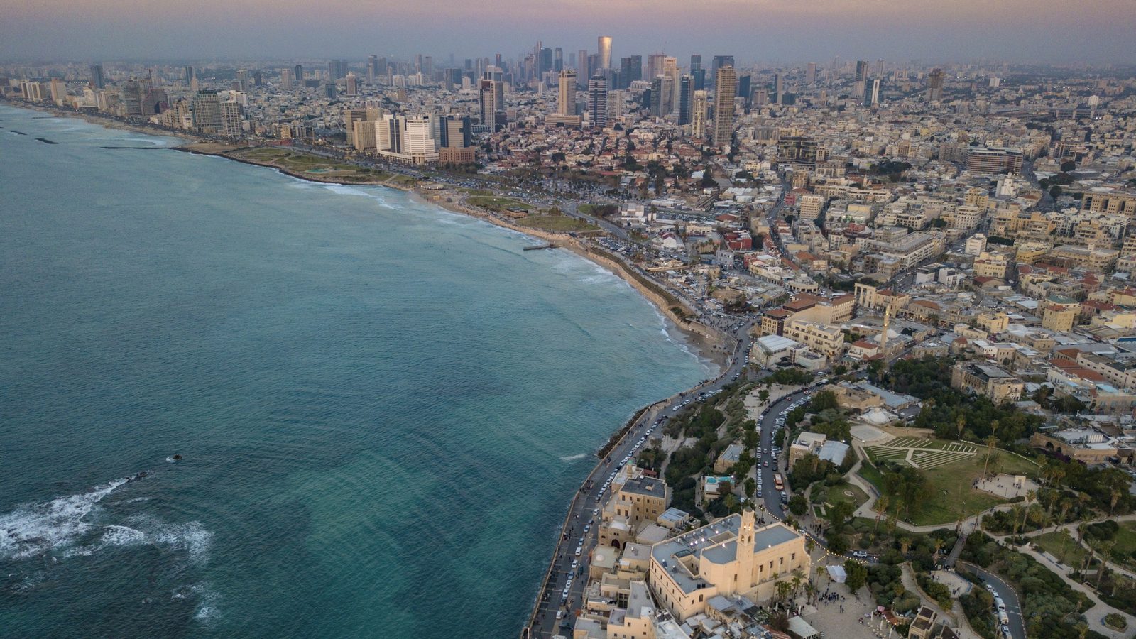 Aerial view of the Tel Aviv coastline at dusk, with old Jaffa and its clock tower church in front and the towers of central Tel Aviv behind