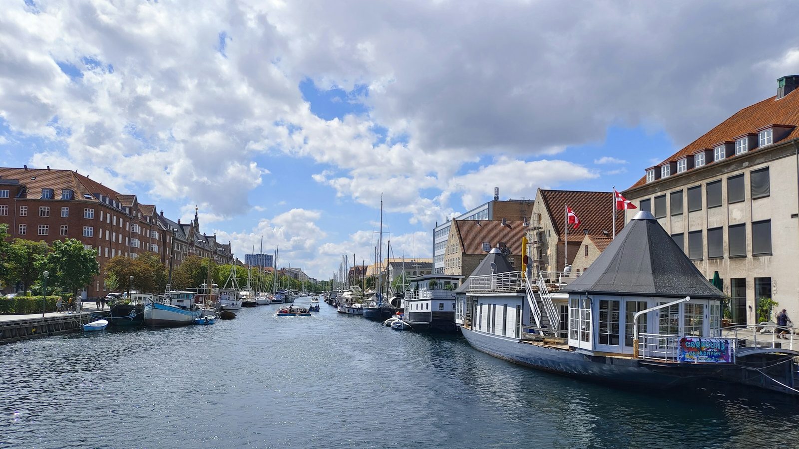 A canal in Copenhagen lined with houseboats and sailing boats, red-brick buildings and Danish flags under a cloudy blue sky