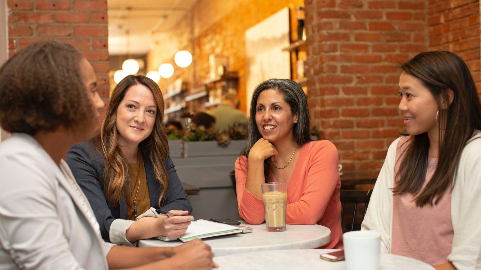 Four women talking around a round marble table in a café with brick walls, one taking notes in a notebook