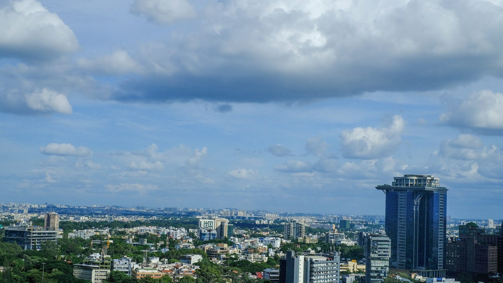 Bengaluru skyline under a cloudy blue sky, with a tall glass tower on the right and green tree cover between low-rise buildings