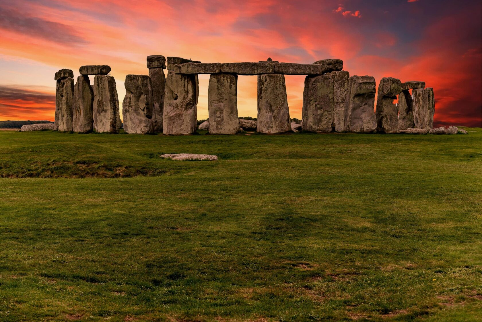 Ancient Stonehenge monument in Wiltshire, UK, with large standing stones arranged in a circle on a grassy field, set against a vibrant orange and pink sunset sky