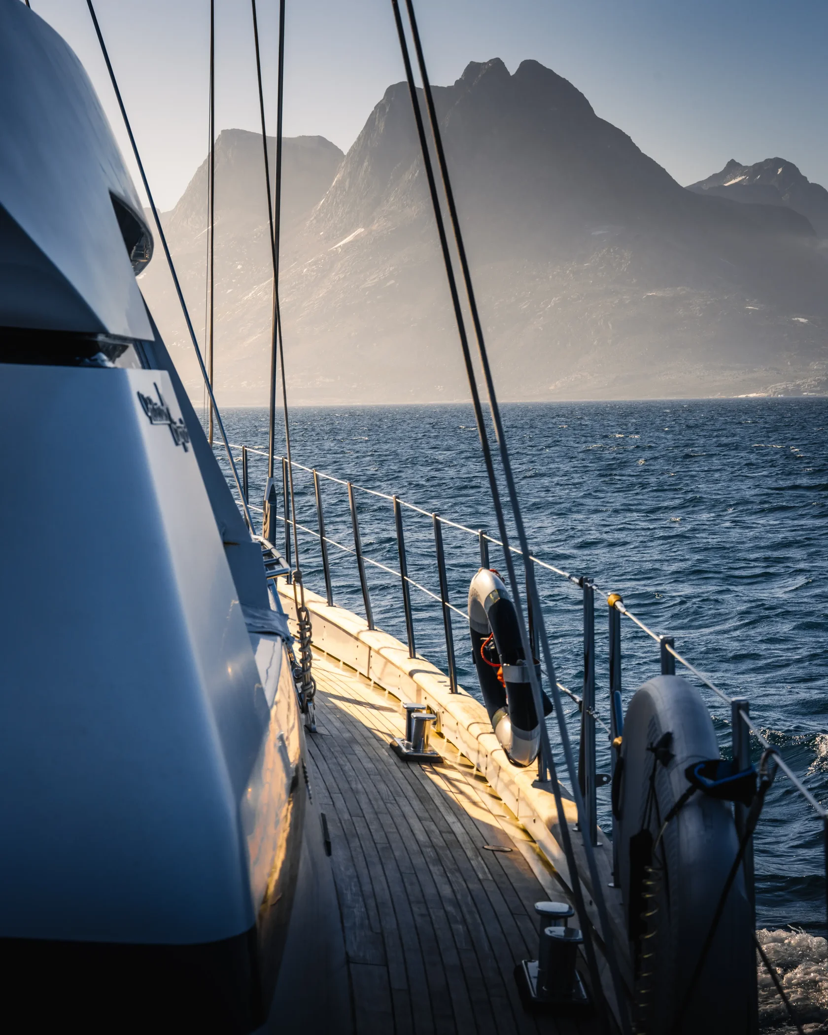 View from the deck of a sailing yacht cruising through the spectacular fjords of East Greenland. Towering Arctic mountains rise above the coastline as golden evening light illuminates the landscape showcasing the unique experience of exploring Greenlands 
