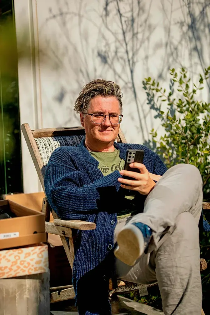 музыкальный продюсер  лейбла REFLEXMUSIC Вячеслав Тюрин сидит в кресле на улице, закинув ногу на ногу, он в очках, волосы развеваются на ветру, он смотрит в телефон