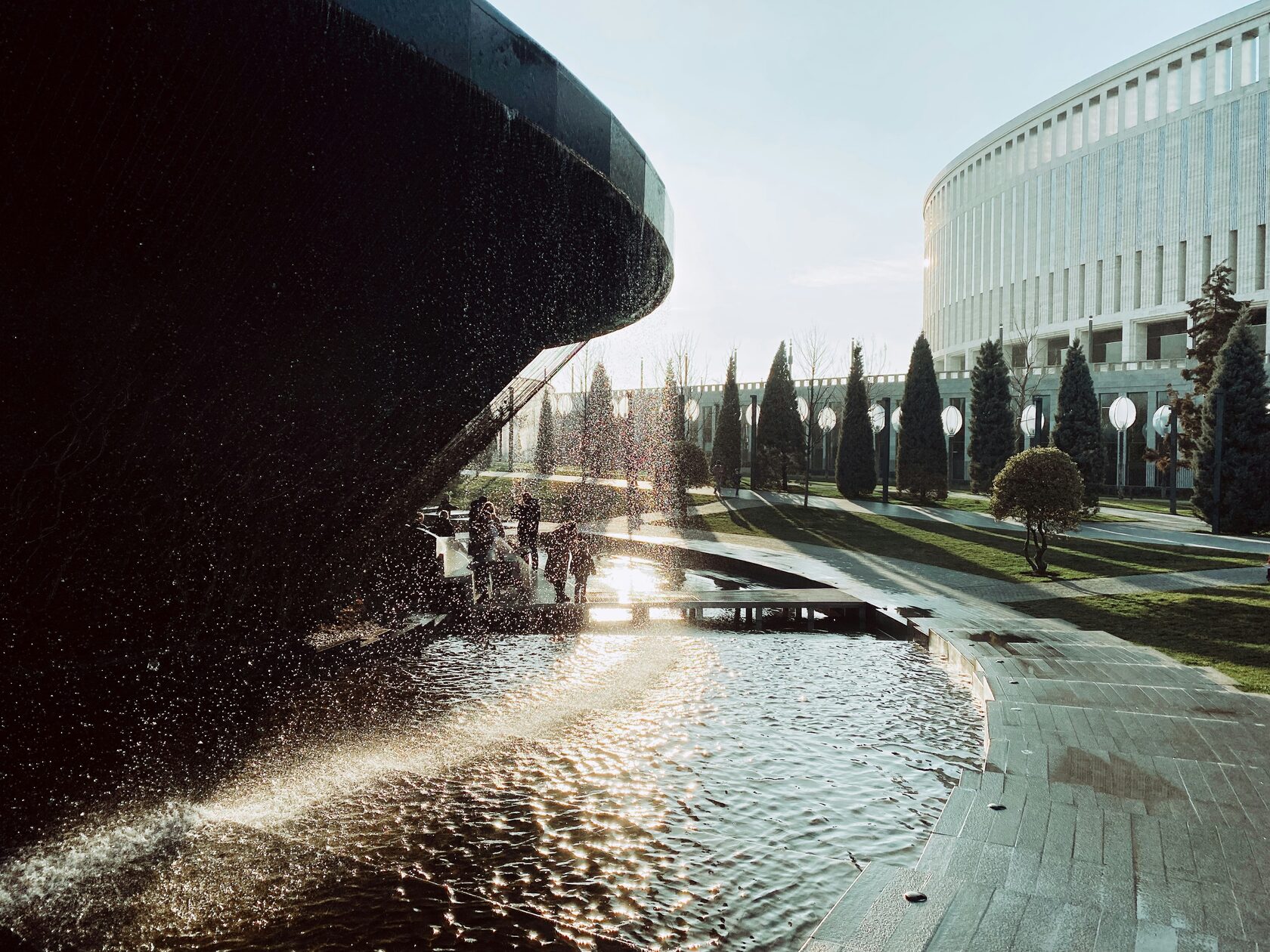 Парк Краснодар: фонтан, прогулочная зона и люди возле современного круглого здания на закате.