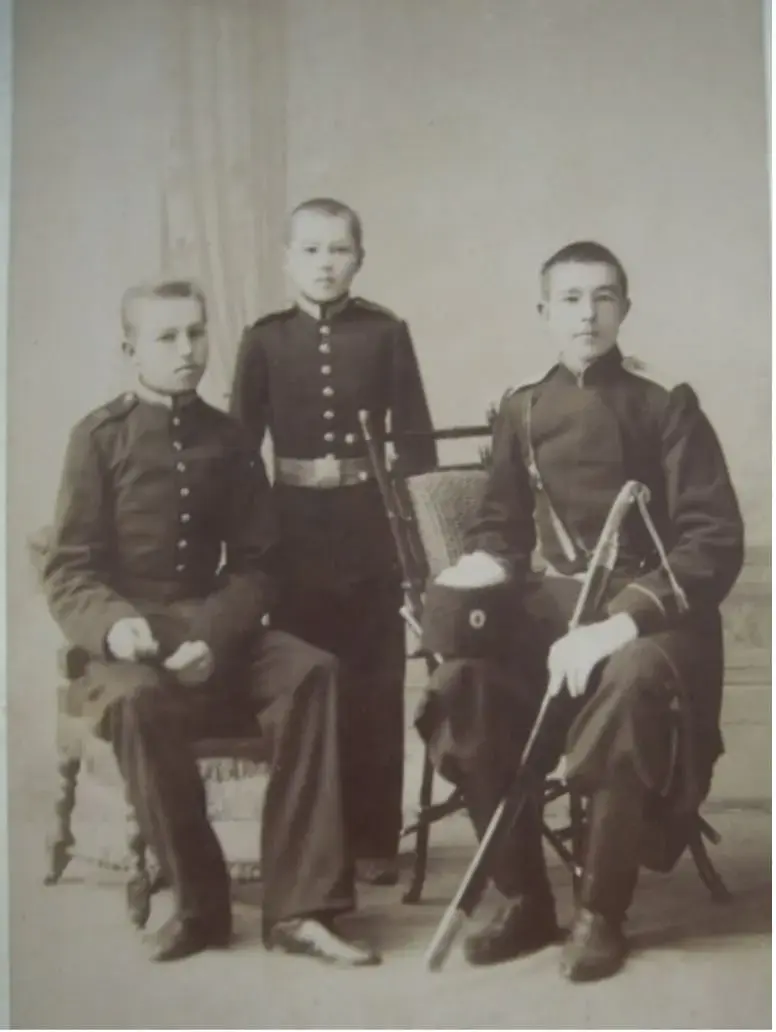 Архивный sepia-снимок конца XIX — начала XX века: трое юношей в кадетской или казачьей форменной одежде позируют в студии; двое сидят на стульях, один стоит между ними, у сидящего справа в руках фуражка и шашка.