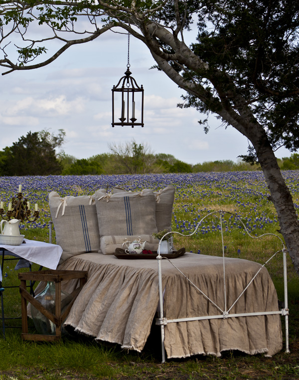 кровать с льняным постельным бельем и сервированным рядом столом возле дерева на лугу с голубыми цветами 