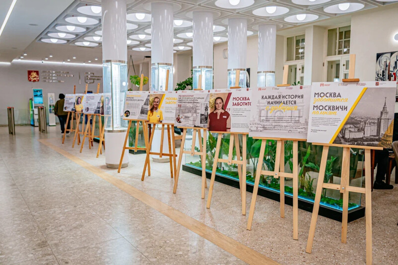 Общий вид фотовыставки «Москва заботится. Москвичи помнят», открывшейся в холле Департамента труда и социальной защиты населения города Москвы ко Дню города. Ряд деревянных мольбертов со стендами о работе социальных работников.