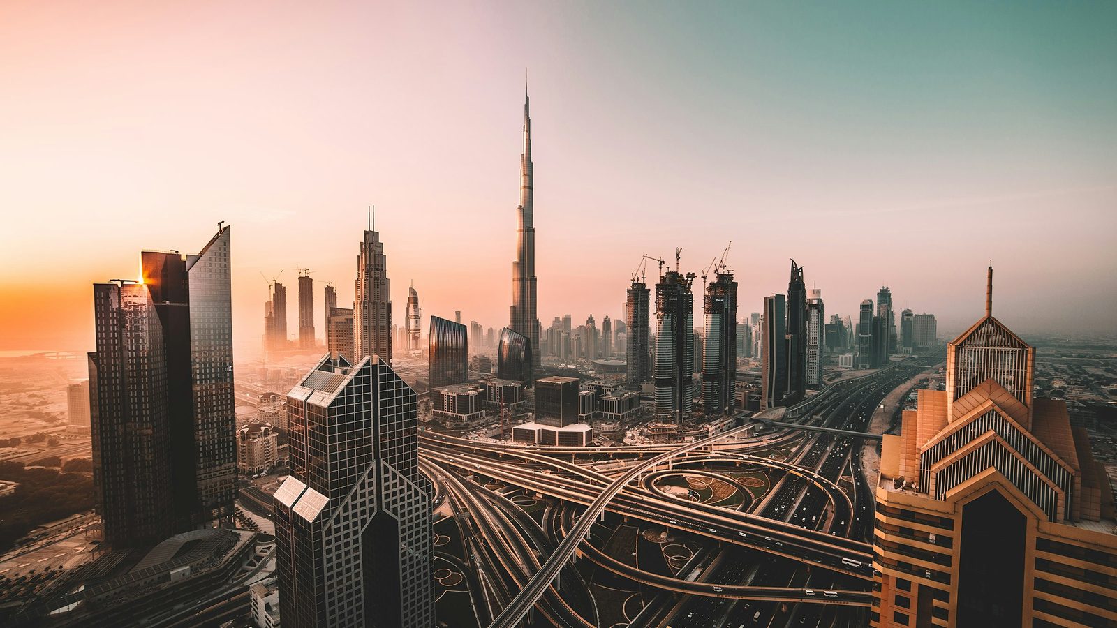 The Burj Khalifa rising above the towers of Downtown Dubai and Sheikh Zayed Road, with a large highway interchange in the foreground, in golden light