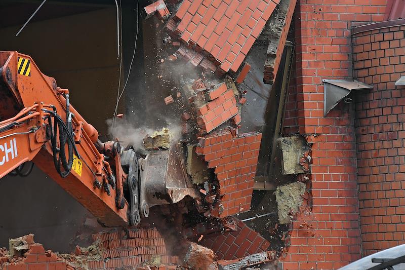 ковш красного экскаватора сносит кирпичную стену