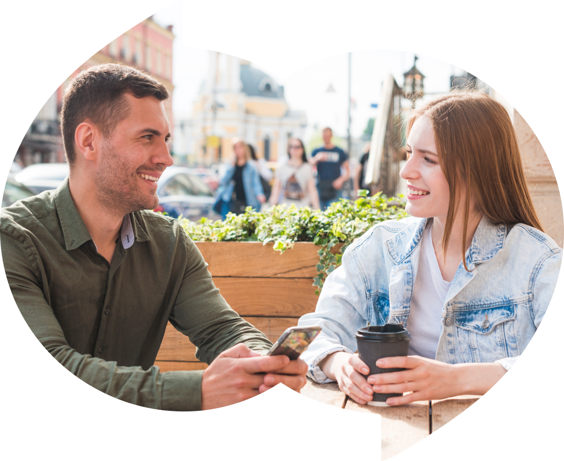 A man and a woman sitting at an outdoor café, smiling and engaging in conversation. The man is holding a smartphone, and the woman has a takeaway coffee cup. They are dressed casually, with the man wearing a green shirt and the woman in a light denim jack