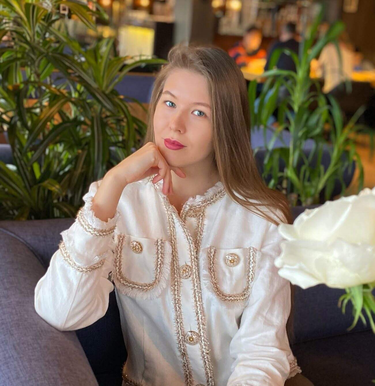 Портретная фотография молодой женщины с длинными прямыми светло-каштановыми волосами, с легким макияжем и красной помадой, в белой куртке-пиджаке с золотистой окантовкой и пуговицами, сидящей на мягком диване и подпирающей подбородок рукой. На заднем плане видны размытые зеленые растения и часть интерьера.