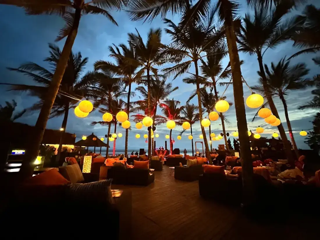 A relaxing evening at a Seminyak beach venue featuring glowing yellow lanterns, palm trees, and comfortable bean bags, showcasing what happens in Bali on Wednesday nights.