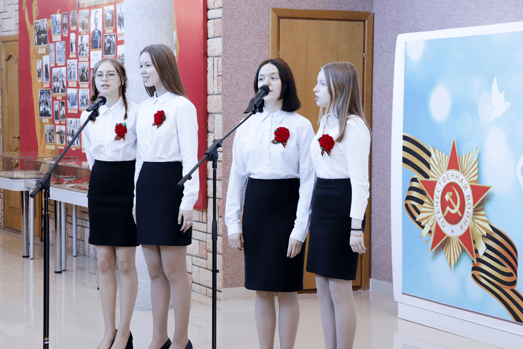 квартет девочек 16 лет в белых блузках и черных юбках-карандашах поет песню перед двумя стойками с микрофонами, девочки стоят по двое