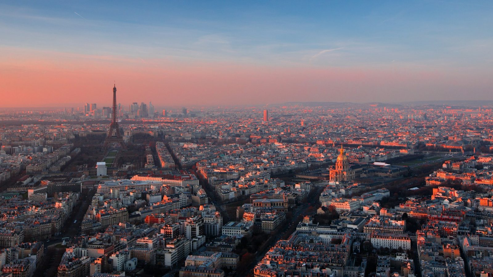 Aerial view of Paris at sunset, with the Eiffel Tower on the left, the towers of La Défense behind it and the golden dome of Les Invalides on the right