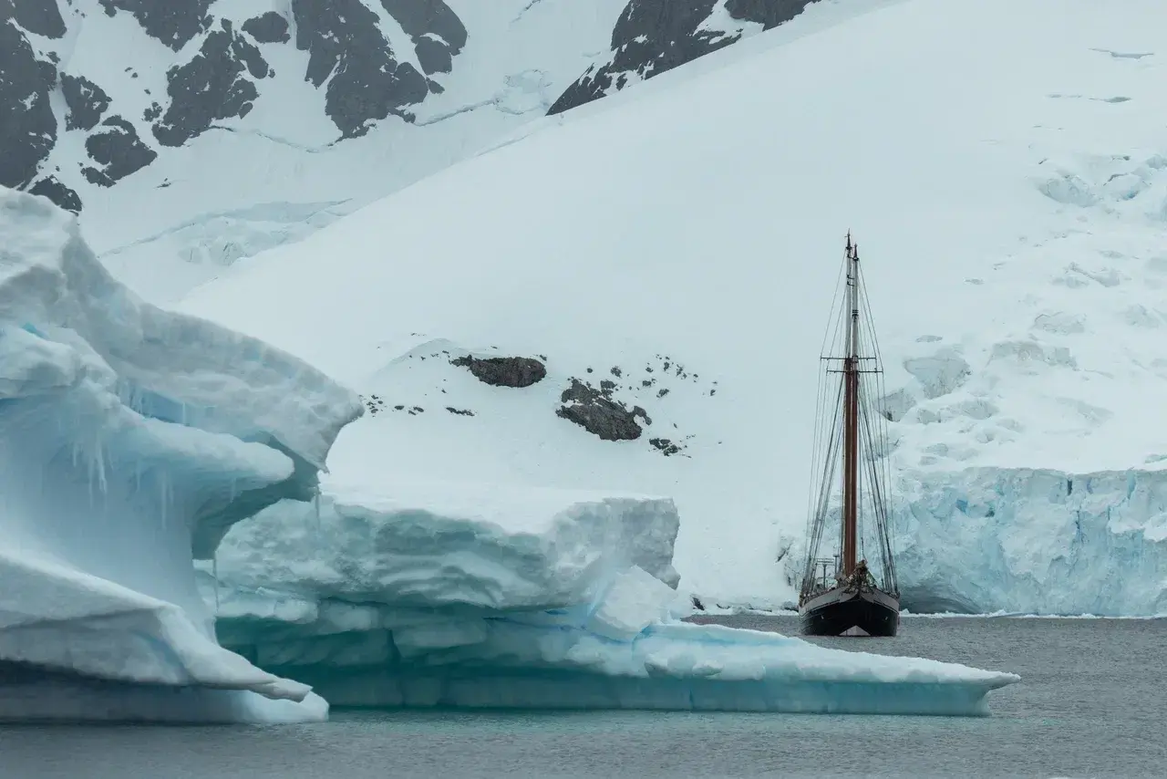 A traditional sailing schooner anchors beside towering icebergs and glaciers in Antarctica. Surrounded by untouched polar landscapes, small expedition yachts can reach remote bays and quiet anchorages that are inaccessible to larger cruise ships, offering a more intimate way to explore the White Continent.