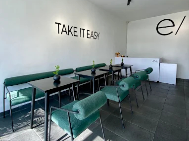 A modern, sunlit cafe interior with the words 'Take It Easy' on the wall, featuring sleek tables and green chairs for a health-conscious take on Russian food in Bali.