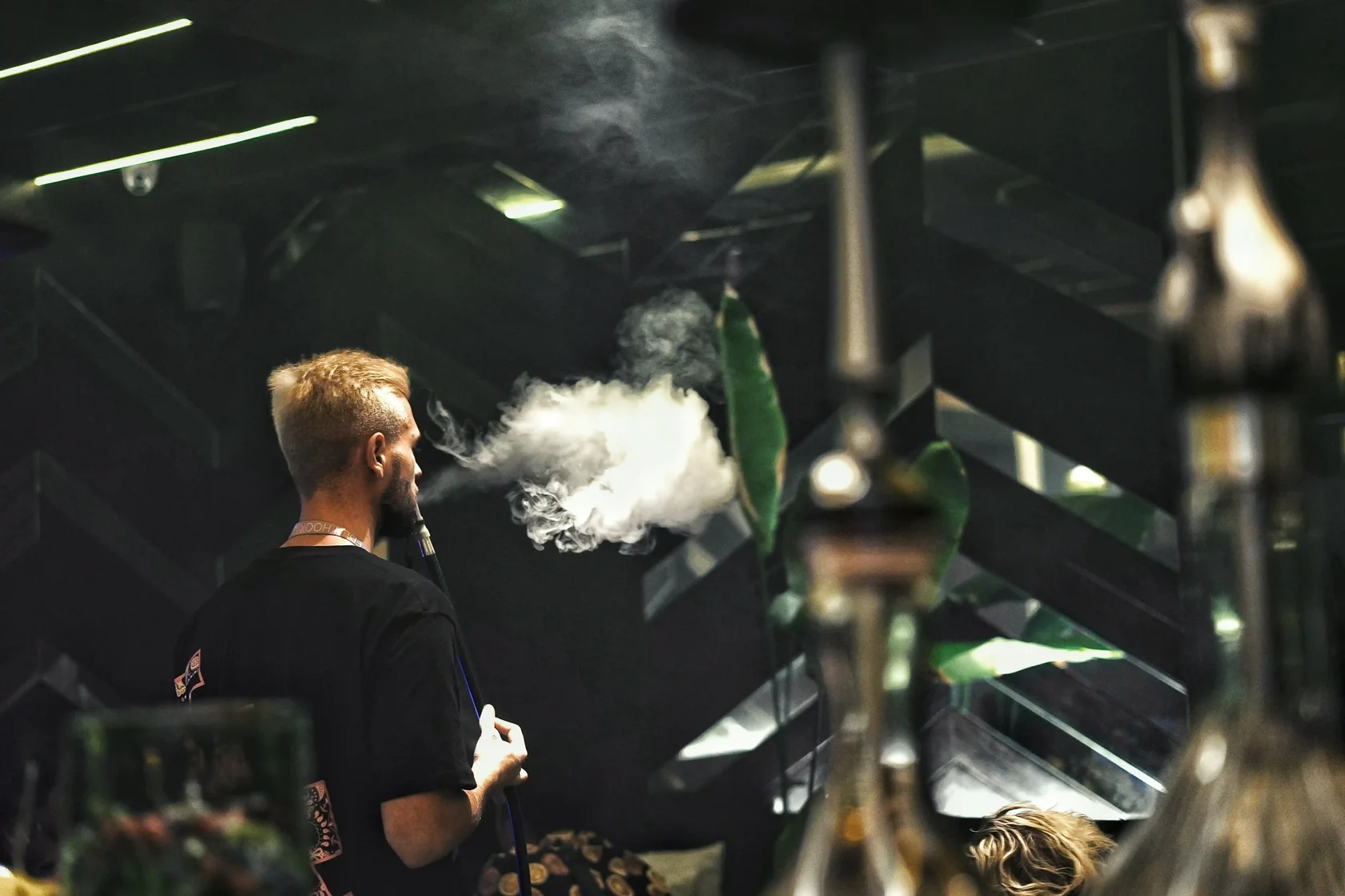 An atmospheric cover photo of a person exhaling a large cloud of smoke from a shisha pipe in a dark, modern lounge, introducing the guide to the top best tobaccos for your shisha in Bali.