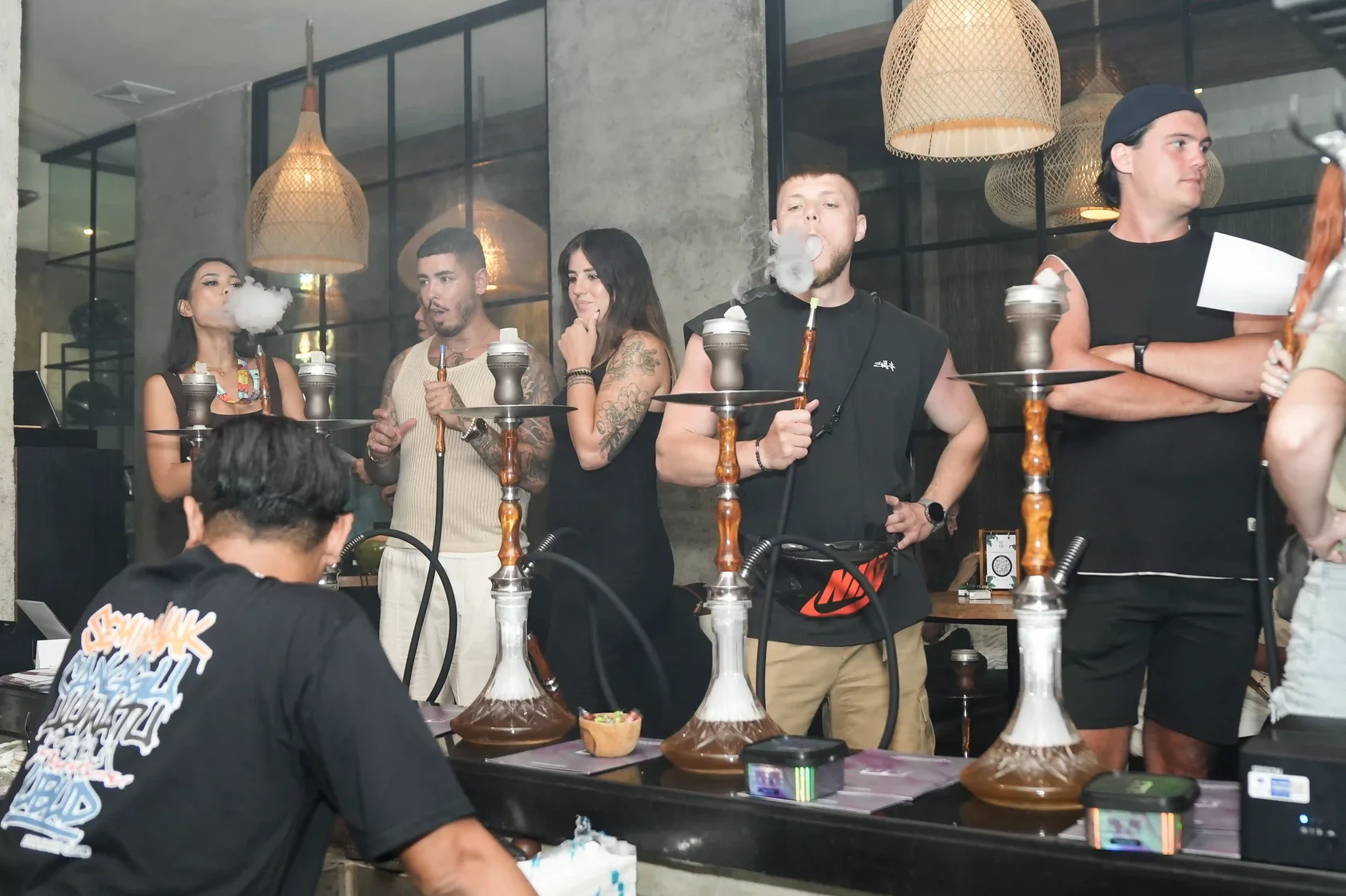 A group of friends gathering at a modern Eden Hookah Club location, featuring unique woven pendant lights and high-quality traditional pipes. Experience championship-level service at one of the best hookah bars in Bali.
