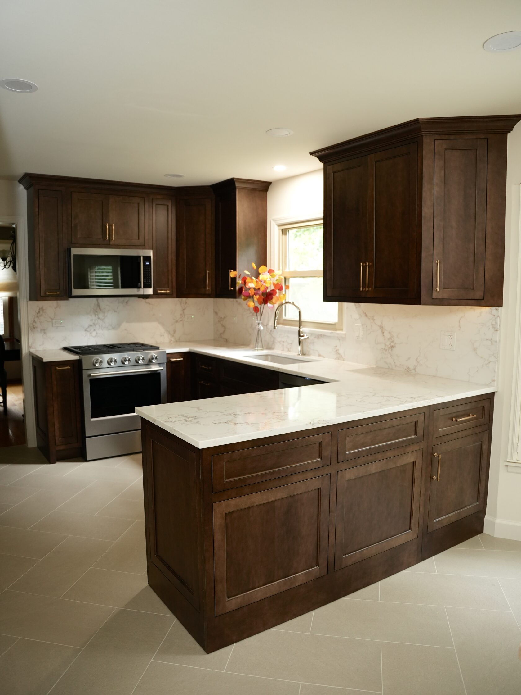 Название: The wider view shows how the dark cabinetry, light surfaces, appliance wall, sink area, and island work together as one coordinated room. - описание: Wide view of a renovated kitchen with dark wood cabinets, stainless steel range and microwave, light counters, a sink below a window, and a large island or peninsula.