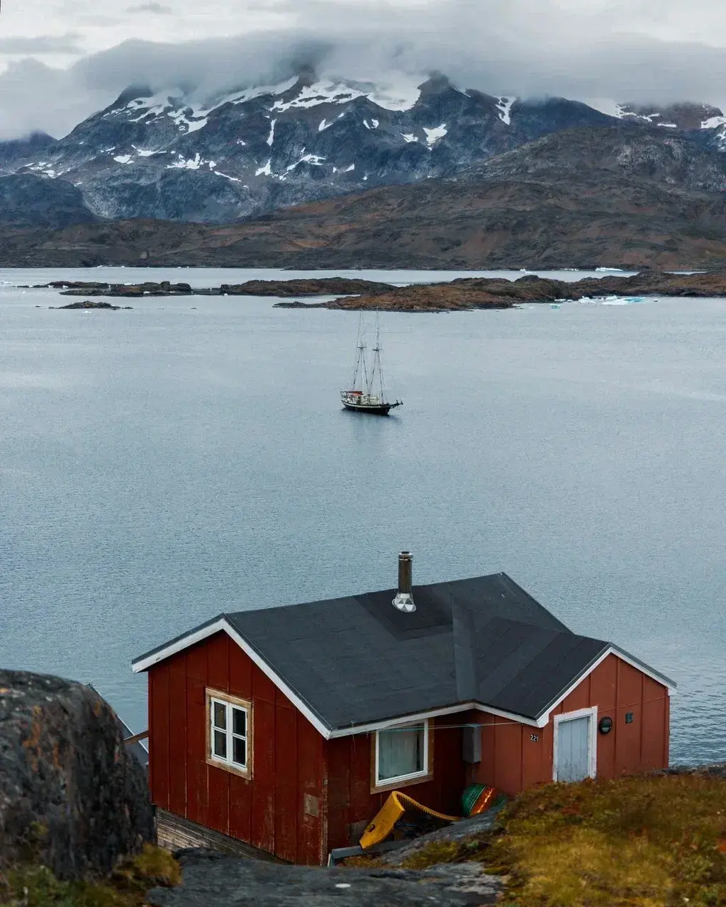 Scenic view of a red house overlooking a calm Greenland fjord with a sailing schooner anchored offshore and snow-covered mountains rising in the background