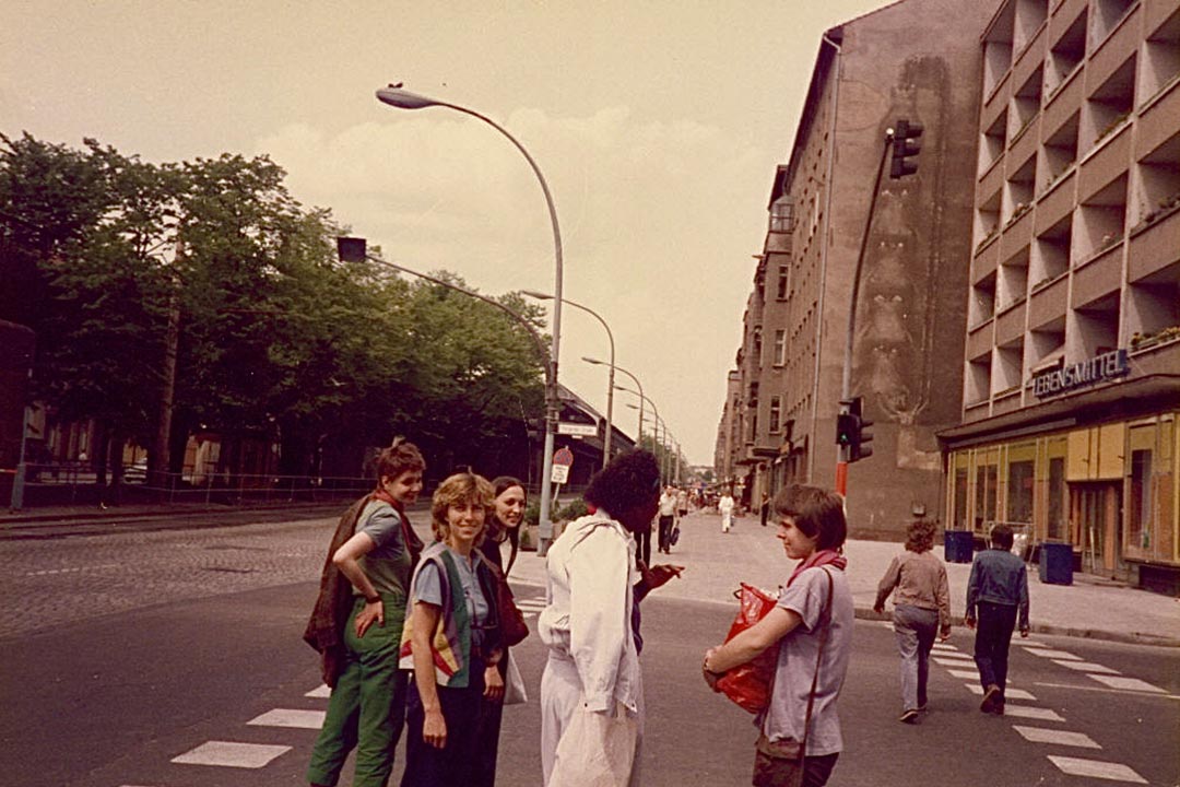 Тифлокомментарий. Цветная фотография. В летний день на пешеходном переходе стоит компания женщин. Это Глория Джозеф и Дагмар Шульц с лесбиянками из ГДР. Женщины разговаривают и улыбаются. Глория одета во все белое, она разговаривает с Дагмар. Дагмар в серой футболке и брюках, у нее короткие русые волосы, она прижимает к груди красную сумку. 
