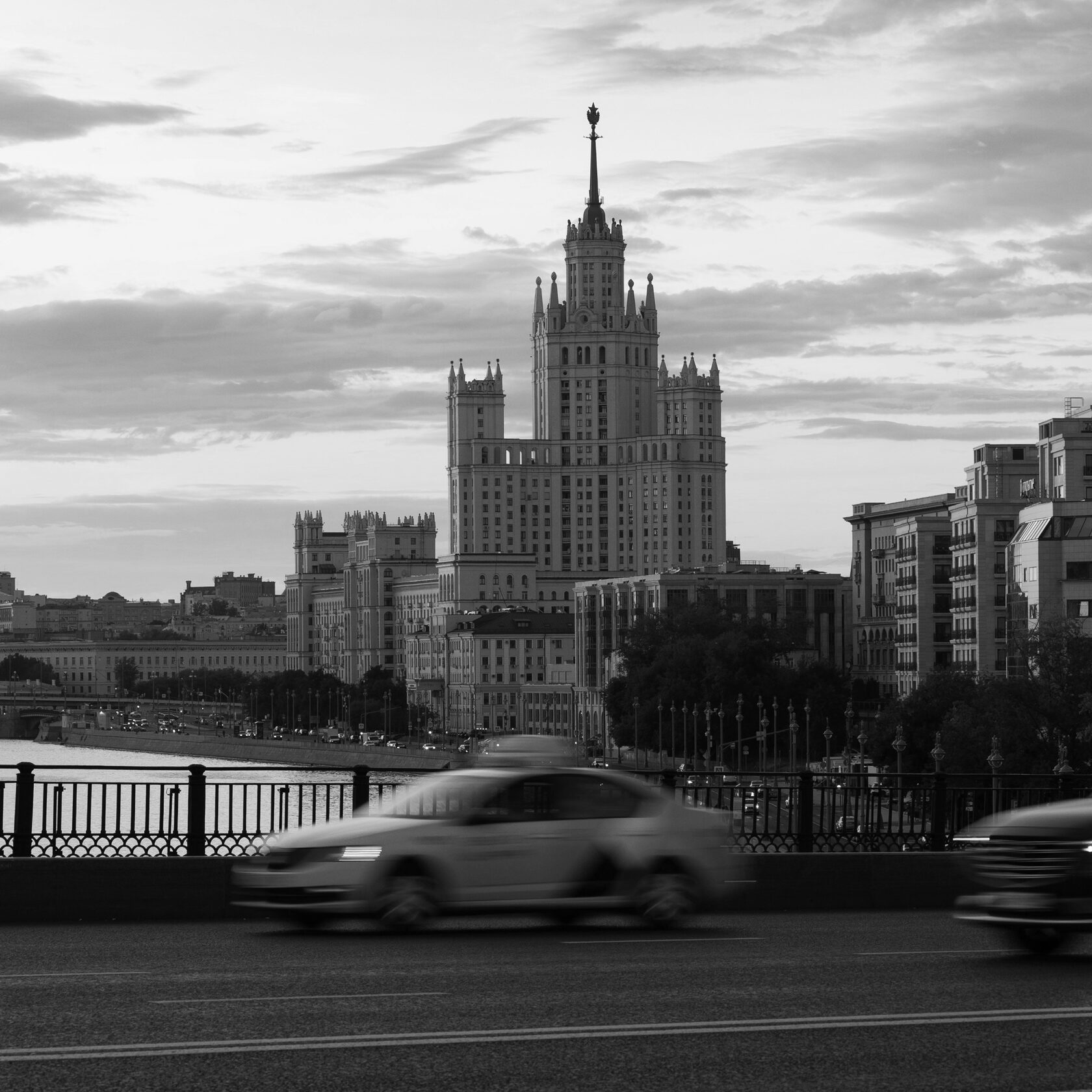 Чёрно-белая фотография гигантской статуи бога Муругана у входа в пещеры Батур (Batu Caves) в Малайзии. На заднем плане видна лестница, ведущая к пещерам, окружённая густой растительностью и скальным массивом. Монументальная фигура статуи доминирует в композиции, подчёркивая религиозное и культурное значение этого места. 