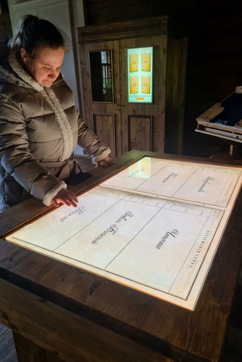 Посетительница в деревянной избе касается подсвеченной витрины-стола, под стеклом — страницы старинной книги; на дверце шкафа позади — небольшой экран