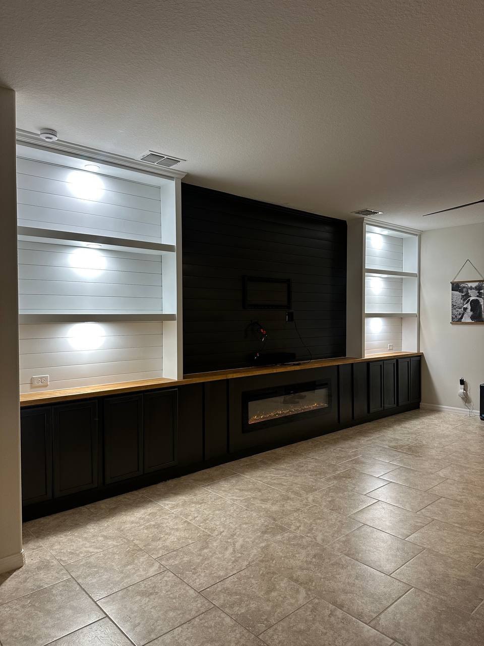 Full view of a custom black shiplap entertainment wall with fireplace, cabinetry, wood countertop, and lighted display shelves