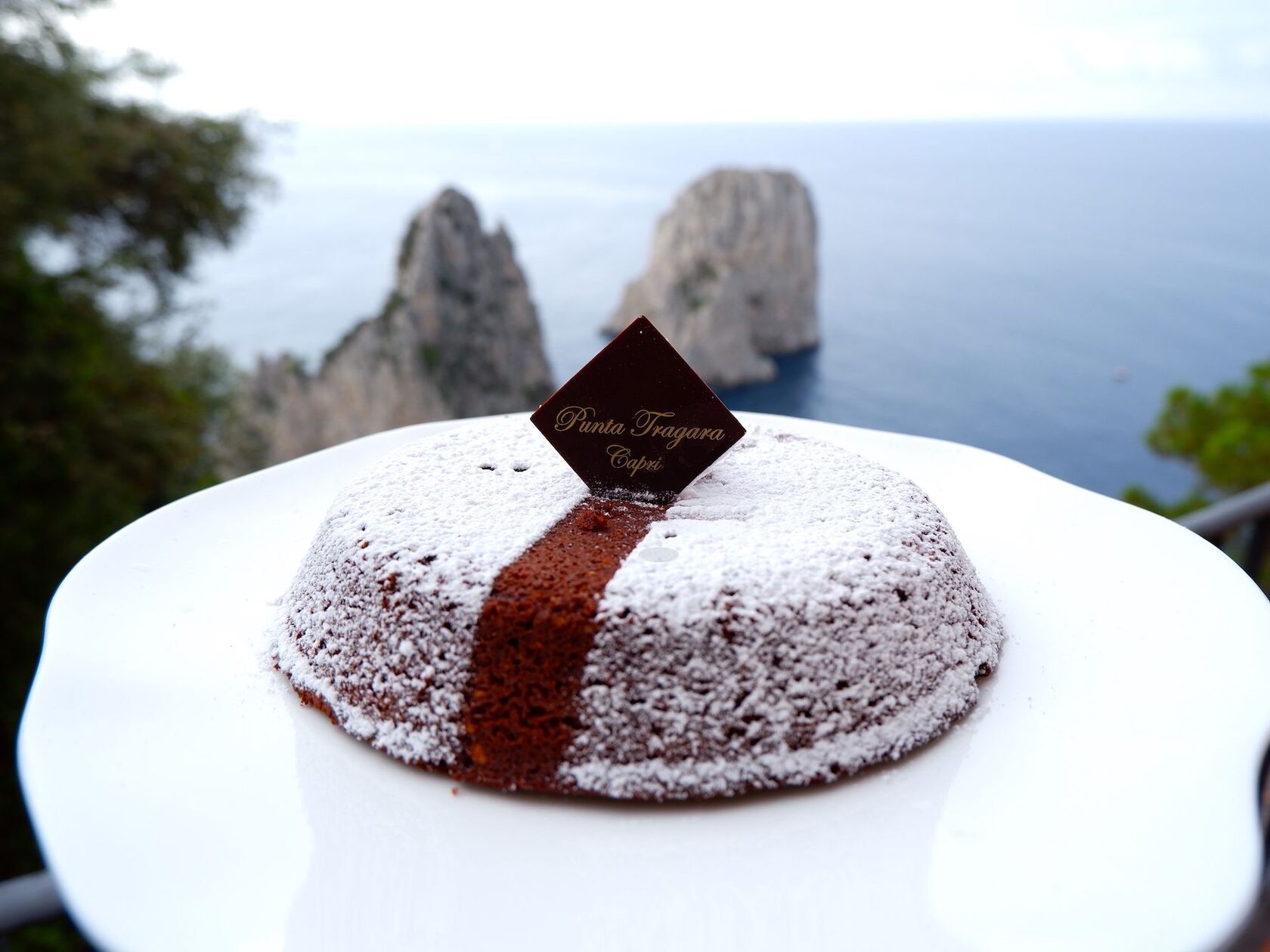 Торт Капрезе с шоколадным логотипом отеля Punta Tragara на фоне моря и знаменитых скал
