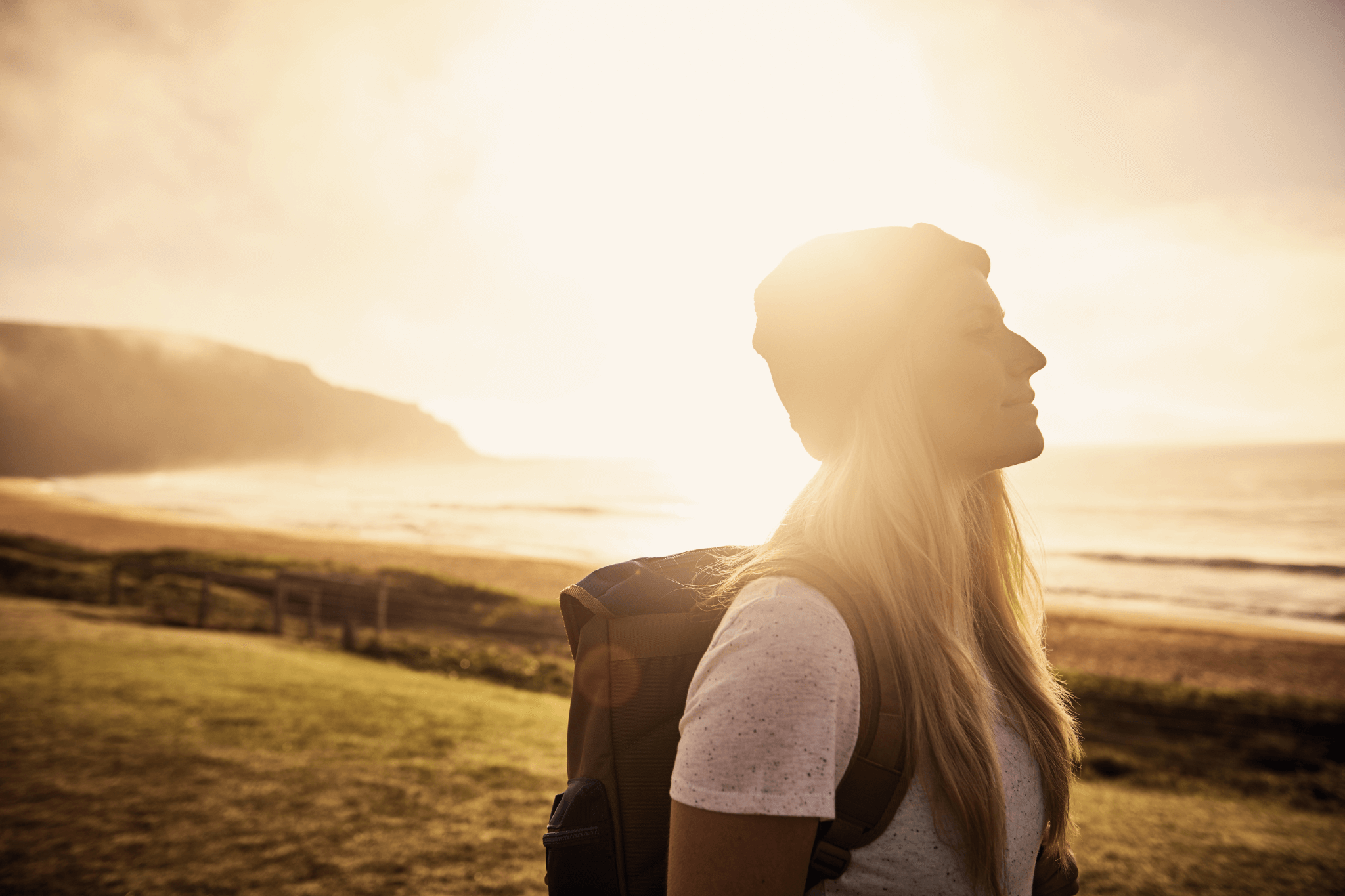Junge Frau mit Rucksack steht entspannt und befreit an Meeresufer bei tiefstehender Sonne