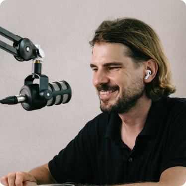 Фотография мужчины со светлыми волнистыми волосами и небольшой бородкой, с улыбкой, в черной футболке, использующего профессиональный микрофон, на заднем плане видна белая стена с размытым фоном.