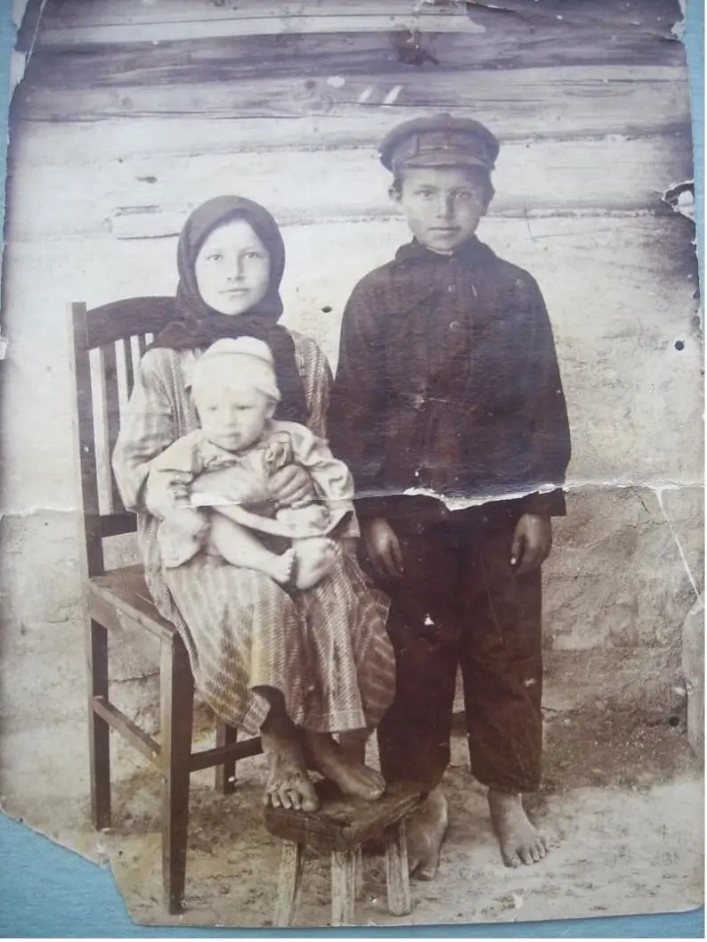 Архивная чёрно-белая фотография начала XX века: босоногая девочка сидит на деревянном стуле, держа на руках младенца, рядом стоит мальчик в фуражке и тёмной одежде. Дети запечатлены на фоне бревенчатой стены сельского дома; снимок сохранил многочисленные следы времени, потертости и повреждения.