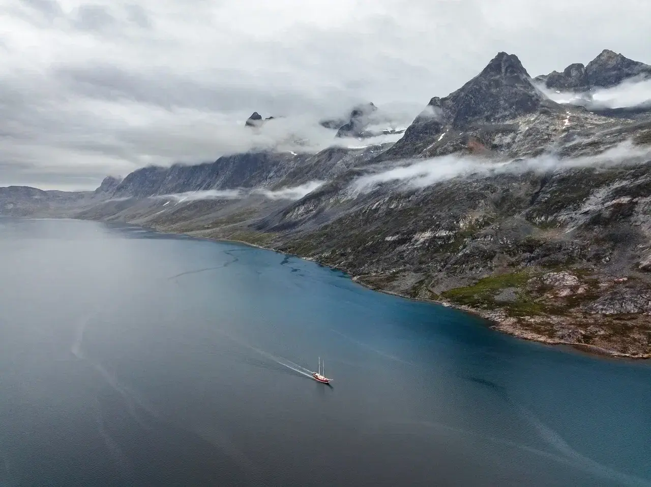 A classic sailing yacht cruising through a remote fjord in East Greenland, surrounded by towering granite peaks, low Arctic clouds and untouched wilderness. This breathtaking landscape showcases the dramatic fjords of Greenland, where small-group sailing expeditions provide close access to pristine nature, glaciers and some of the most spectacular scenery in the Arctic