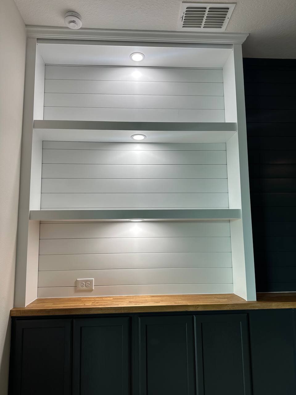 Custom white display shelving with shiplap backing, crown molding, recessed lighting, and black lower cabinets