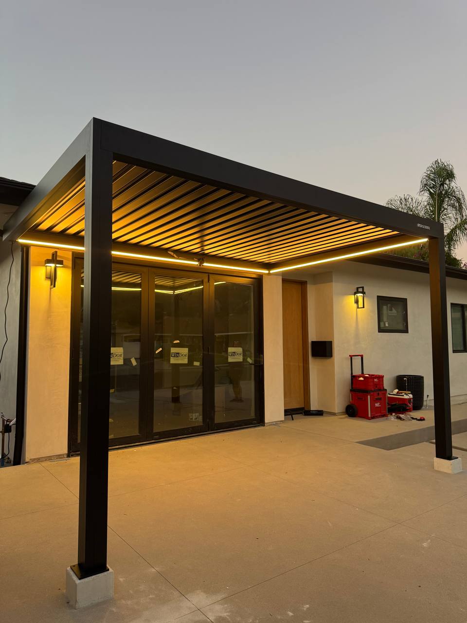 Warm perimeter lighting beneath the black louvered pergola outside the house.
