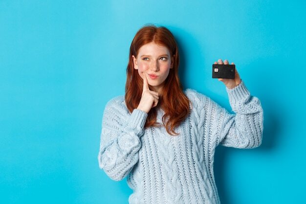 Image of thoughtful redhead girl thinking about shopping, showing credit card and pondering, standing over blue background