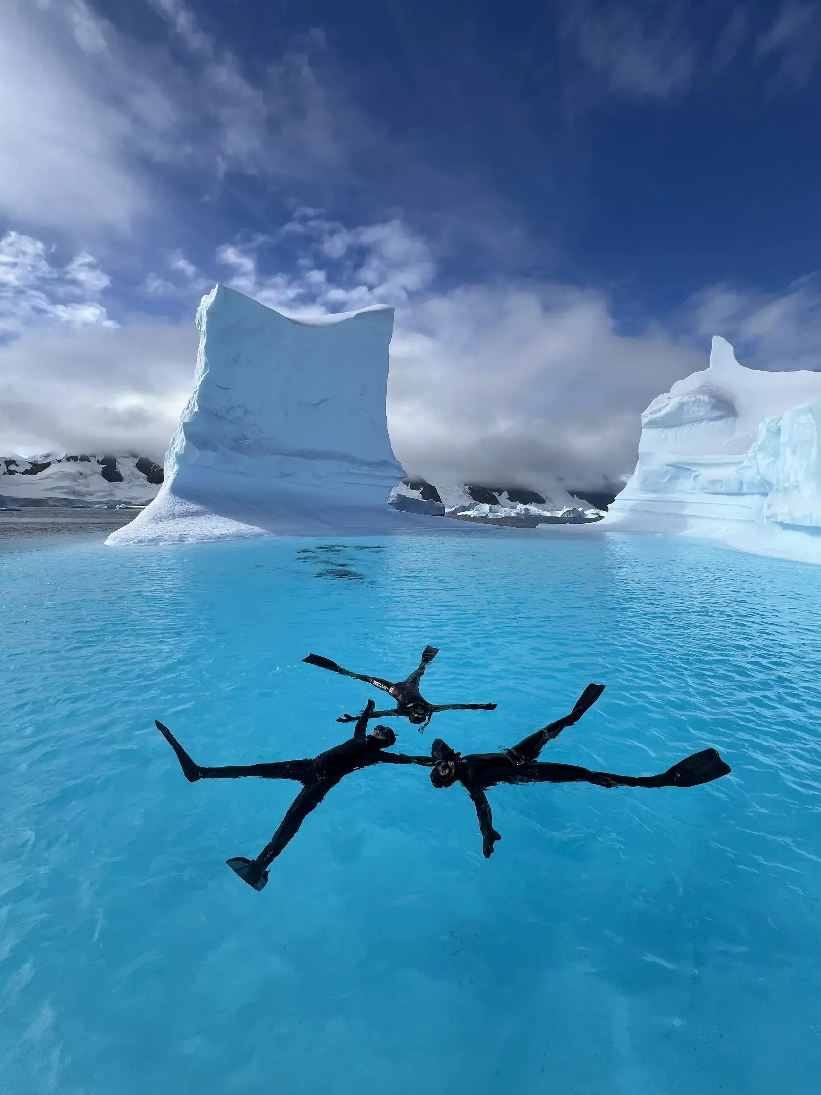 Freedivers exploring crystal-clear Antarctic waters among towering icebergs during a polar freediving expedition accessible by expedition sailing yacht.