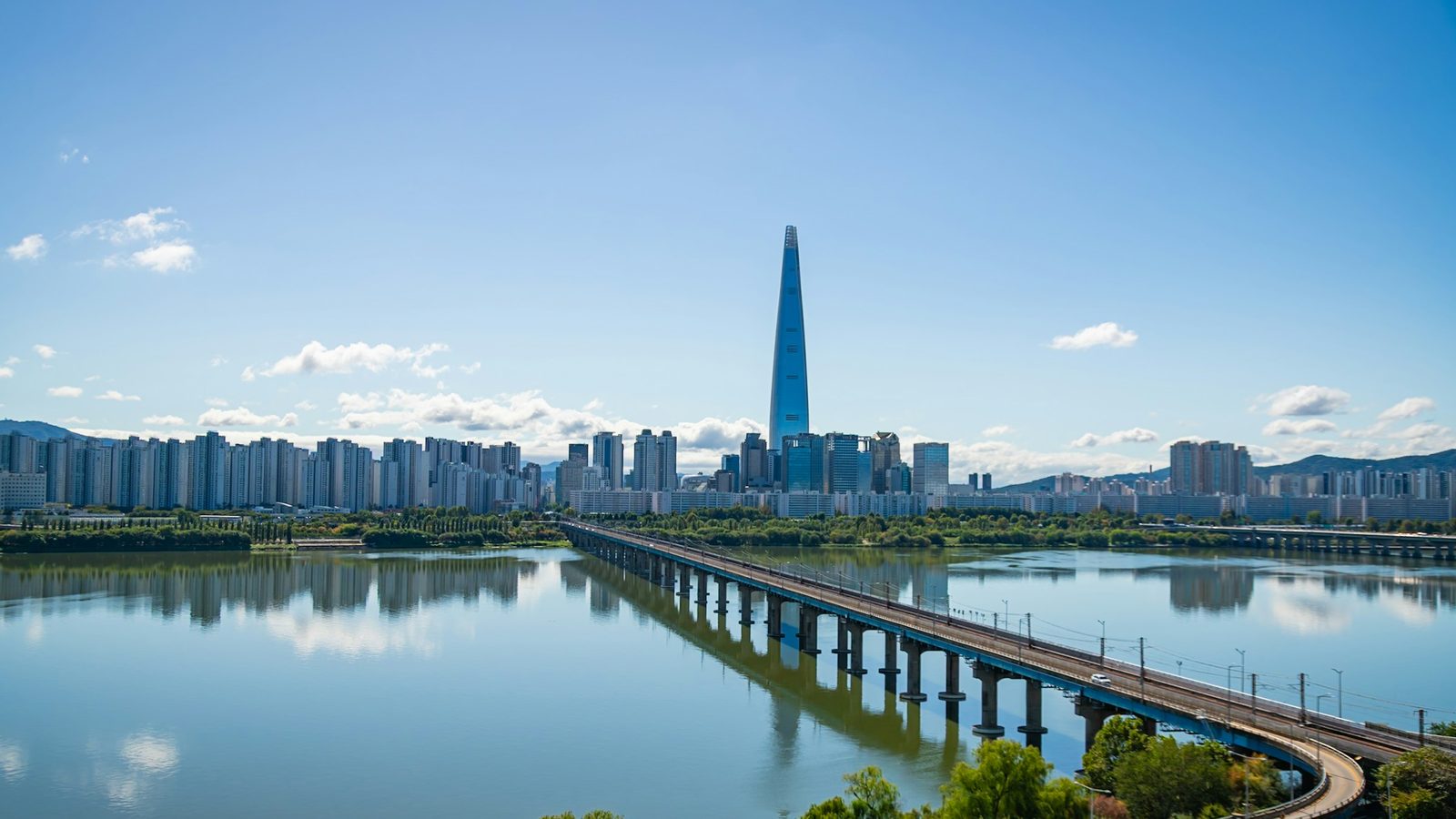 Seoul skyline across the Han River on a clear day, with the Lotte World Tower rising above apartment blocks and a long railway bridge in the foreground