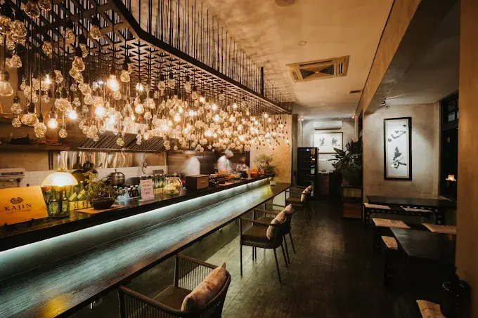 The stylish and moody atmosphere inside Kajin, with a long wooden sushi bar illuminated by many hanging Edison bulbs. Perfect for a contemporary sushi date night in Seminyak.