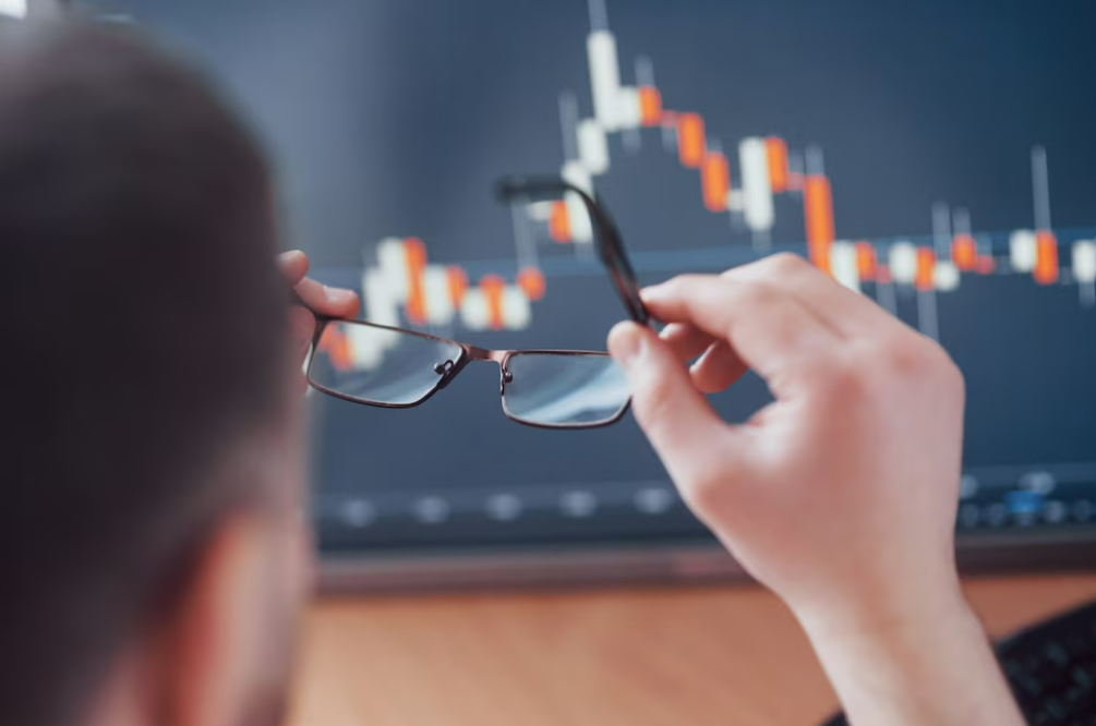Hombre de espaldas sostiene sus gafas, mientras analiza una pantalla con gráficos de precios para aprender trading