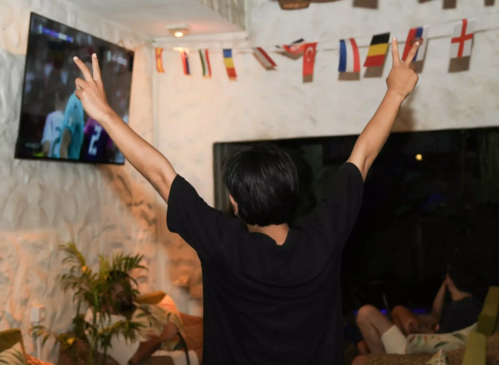 A fan’s perspective from behind, celebrating with hands raised high making victory signs while watching an exciting football match on a large screen in a decorated venue. The energy here makes this the best event in Bali for major sporting finals.