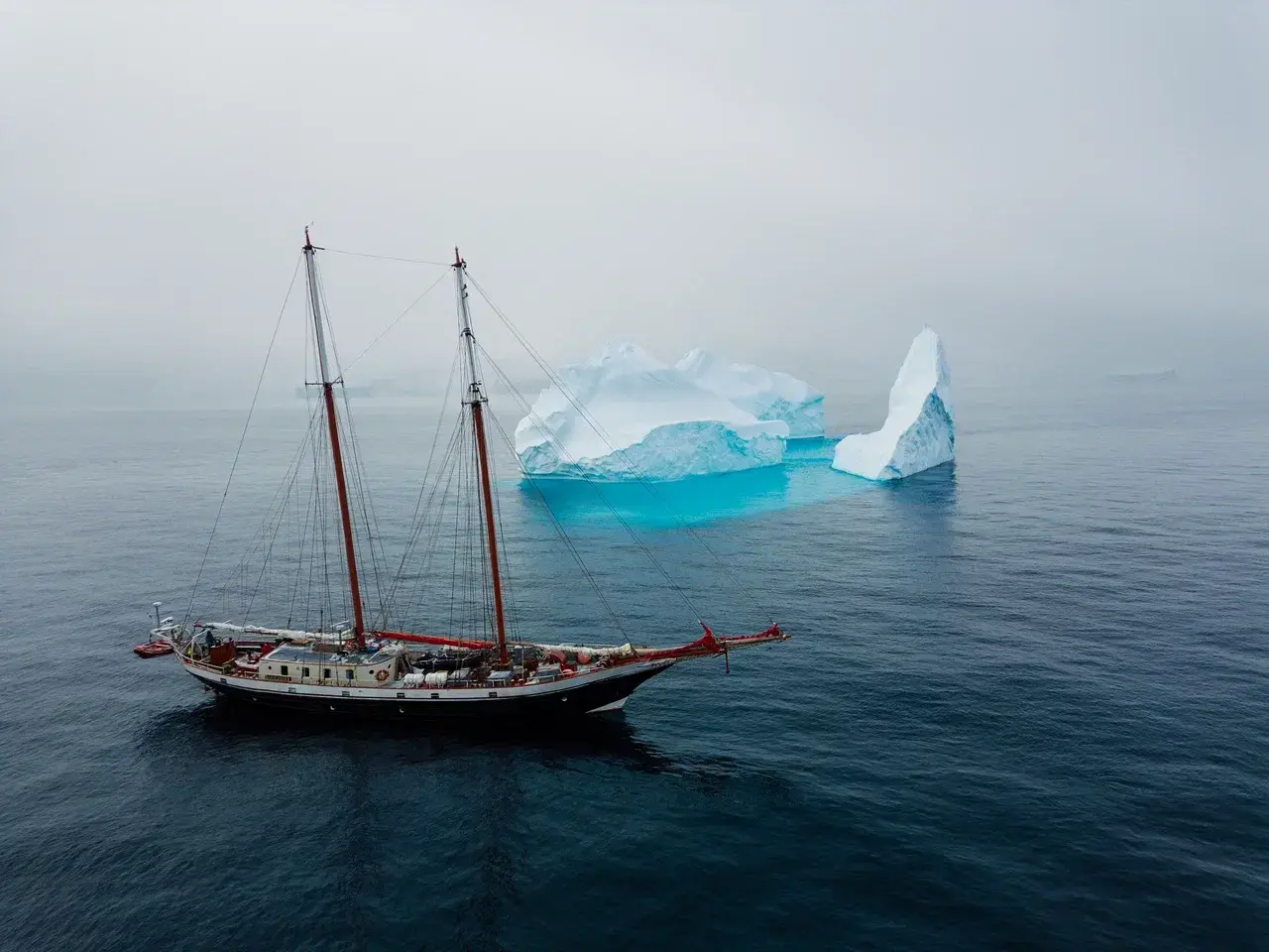 The schooner Amazone sailing through remote polar waters beside a bright blue iceberg. Designed for small-group expeditions, Amazone takes travelers deep into the spectacular landscapes of Antarctica and the Arctic, combining classic sailing with close-up exploration of icebergs, glaciers and wildlife.