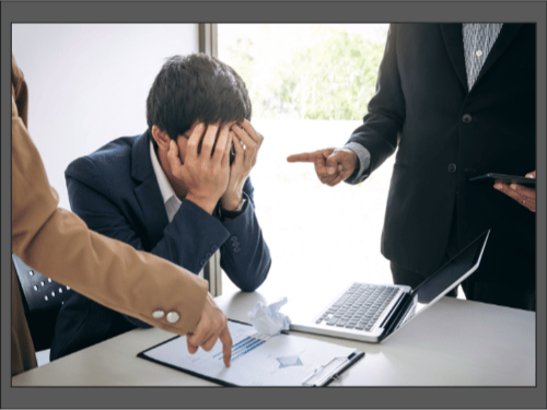 Мужчина в костюме сидит за столом, закрыв лицо руками, коллеги указывают на него пальцами и бумагами.